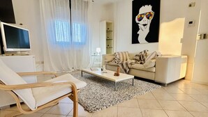 A modern living room with a white sofa, a wooden chair, a coffee table, and a TV. - Veliero Flexrent - Abyssinia Real Estate (Riccione)