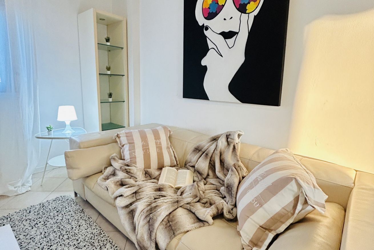 A cozy living room with a beige sofa, striped pillows, and a fur throw. There's a glass cabinet with plants and a framed artwork on the wall.