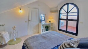 A bedroom with a large arched window, a bed with patterned bedding, a bedside table with a lamp, and a small plant. - Veliero Flexrent - Abyssinia Real Estate (Riccione)