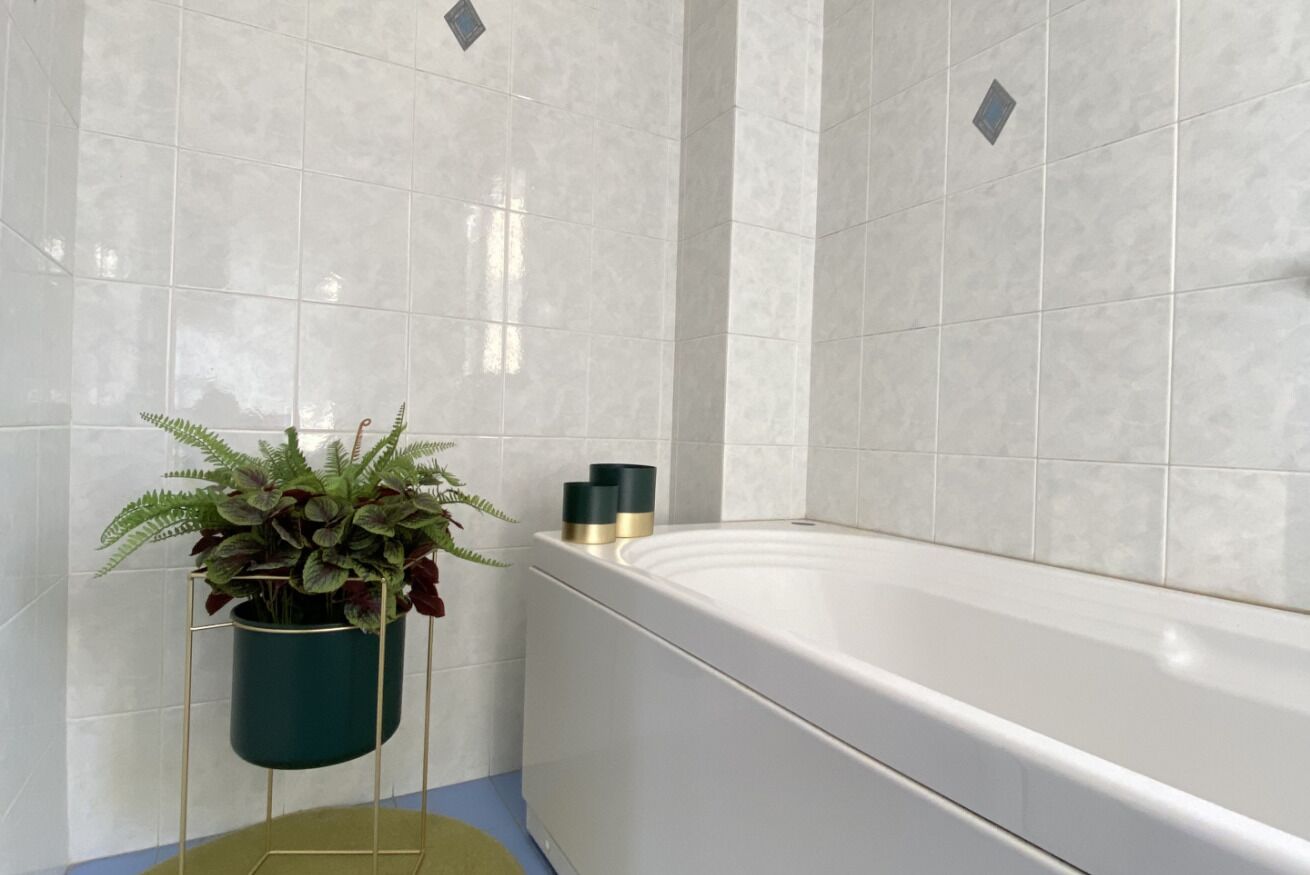 Bagno con piastrelle bianche, una vasca da bagno, una pianta verde su un supporto in metallo e due flaconi neri e dorati su un lato.