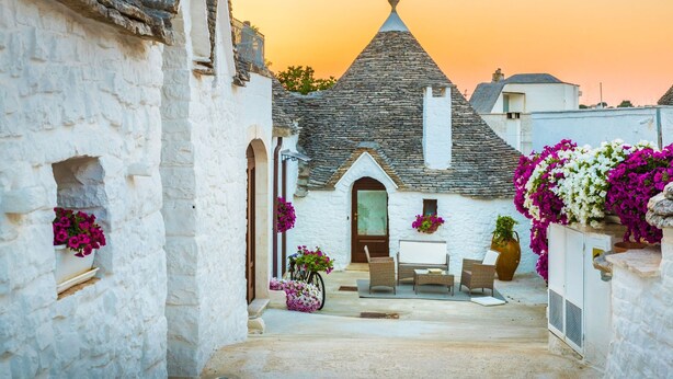 A narrow street with a white building featuring a stone roof and a small porch with a table and chairs.