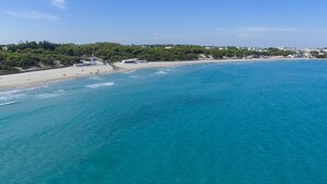 A coastal beach with clear turquoise waters, a sandy shore, and buildings in the background.