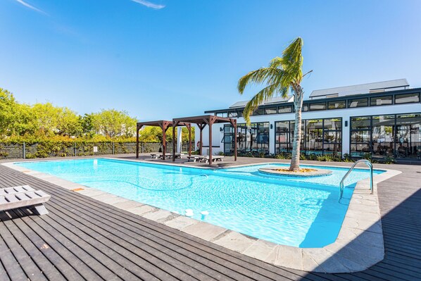 A modern outdoor swimming pool with a wooden deck, a covered seating area, and a palm tree. - The Sandown 178 by CTHA (Cape Town)
