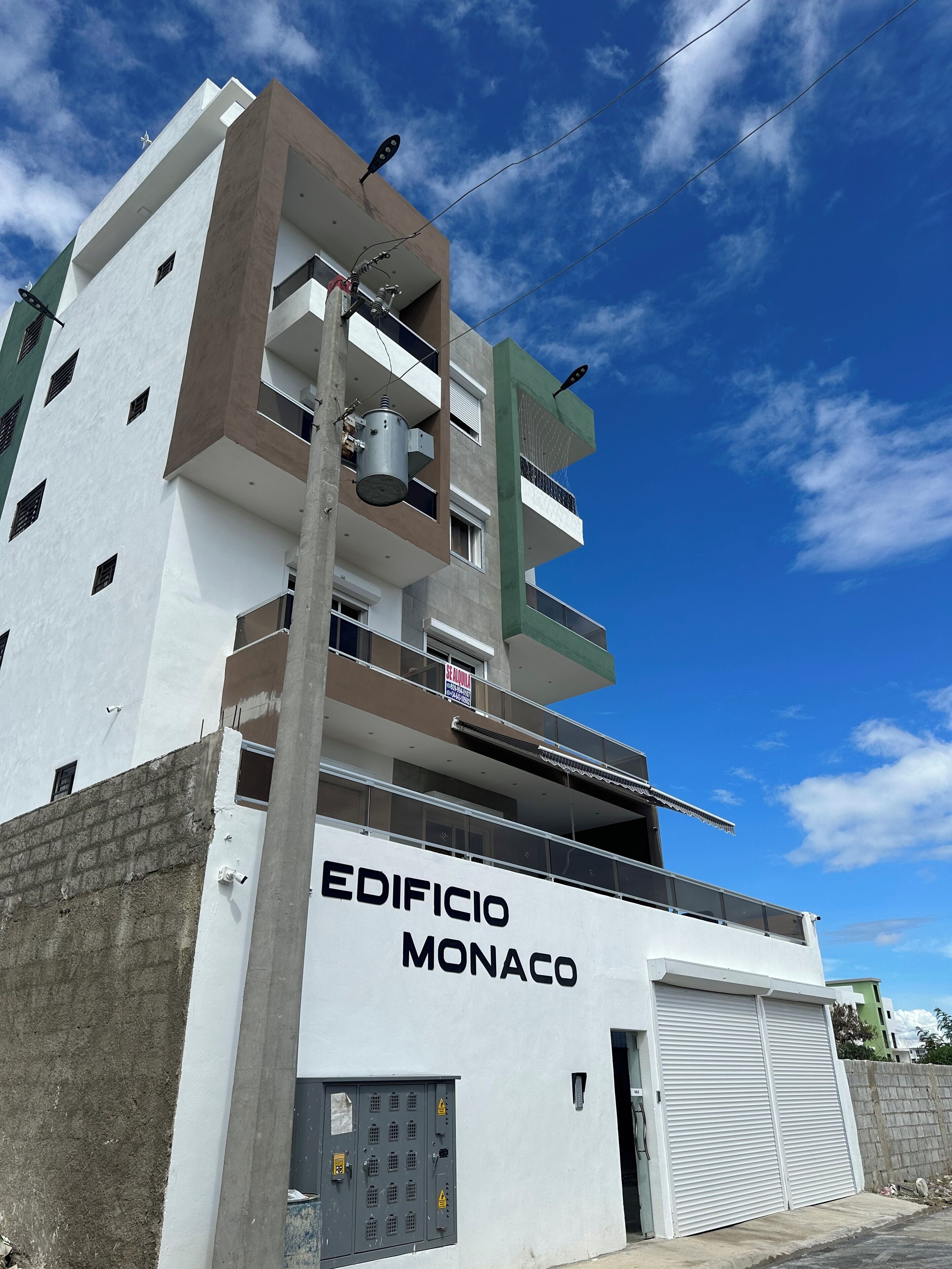A multi-story building with the name "EDIFICIO MONACO" on the facade, featuring balconies, a garage, and visible electrical panels.