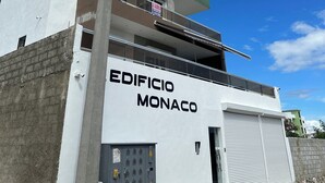 Un edificio de varios pisos con el nombre "EDIFICIO MONACO" en la fachada, que cuenta con balcones, un garaje y paneles eléctricos visibles.