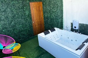 Ein weißes Whirlpool mit mehreren Düsen, eine Holztür und bunte Stühle in einem Gartenbereich mit künstlichem Rasen.