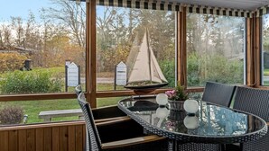 Een overdekt terras met een ronde glazen tafel, rieten stoelen en een model zeilboot.