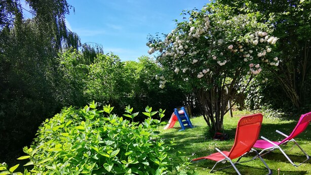 Un jardín con un tobogán, sillas rojas y abundante vegetación.