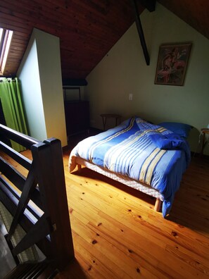 Une chambre avec un plancher en bois, un lit recouvert d’une couvre-lit à rayures, un tableau accroché au mur et une fenêtre avec des rideaux.