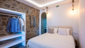 Une chambre avec un mur en pierre, un lit avec une literie blanche, un luminaire suspendu et une armoire remplie de vêtements.