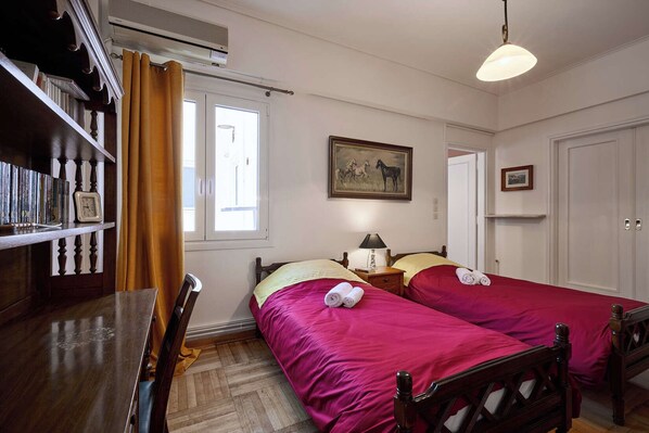 Una habitación con dos camas, un escritorio de madera, una estantería, un cuadro y una ventana con cortinas.
