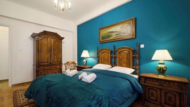 Een slaapkamer met een groot bed, houten hoofdbord, twee nachtkastjes met lampen, een houten kledingkast en een schilderij aan de muur.