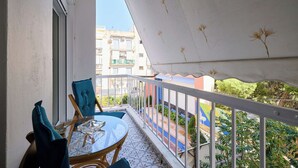 Een balkon met een tafel en stoelen, een wit zonnescherm en uitzicht op een kleurrijk gebouw en groen.