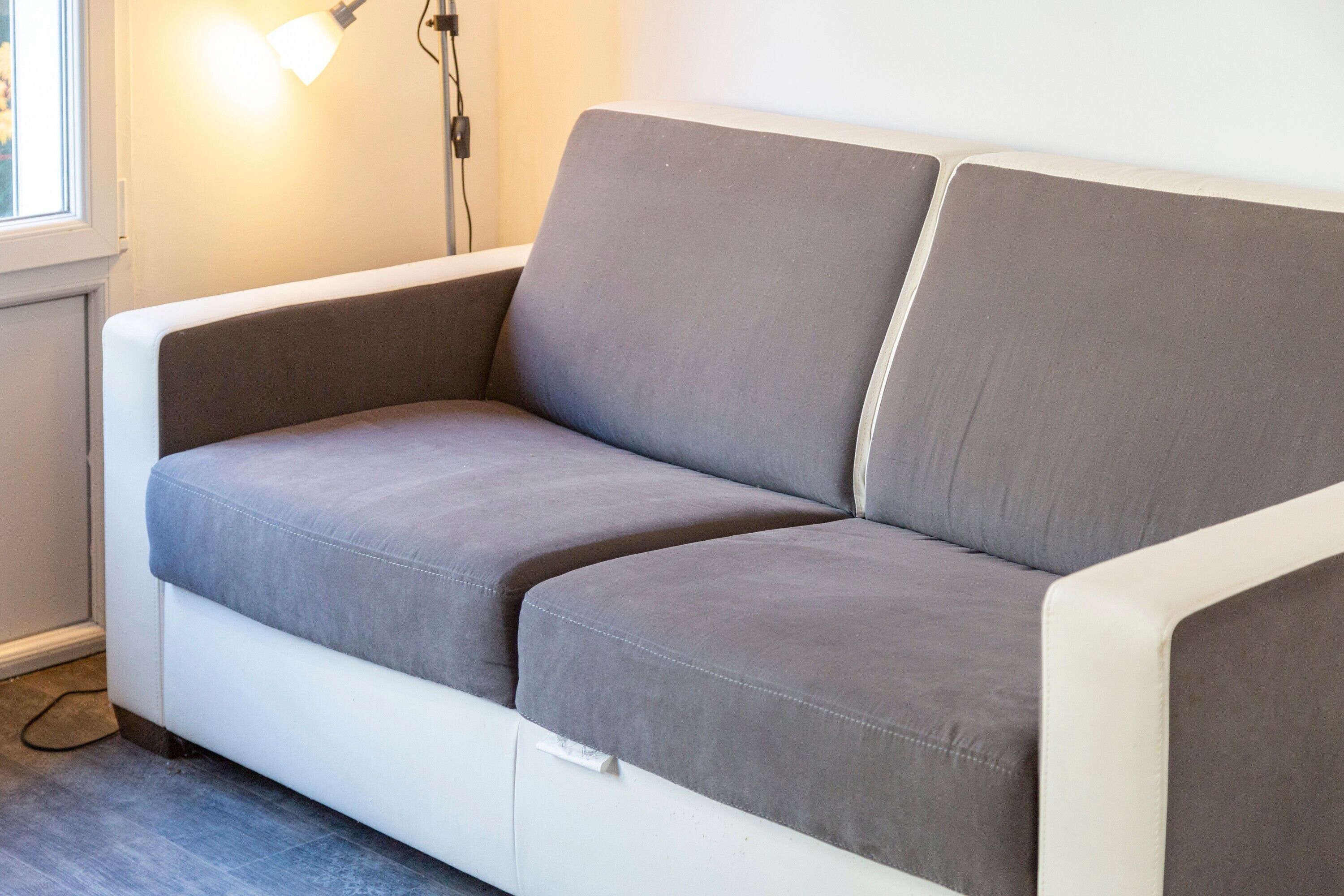 A white sofa with grey cushions in a room with a window and a wall-mounted lamp.