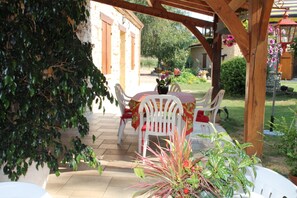 Ein überdachter Patio-Bereich mit Tisch und Stühlen, umgeben von viel Grün und einem Steinhaus.