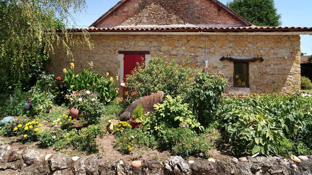 A stone building with a red door, surrounded by a garden with various plants and flowers.