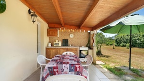 A covered patio with a table and chairs, a wooden ceiling, a kitchen area, and a green umbrella.