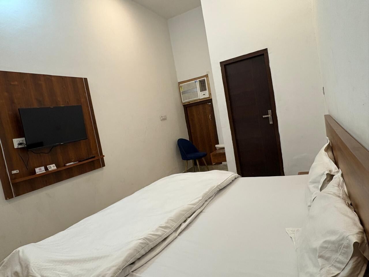 A single-bed room with a wooden headboard, a mounted TV, a blue chair, and a door.