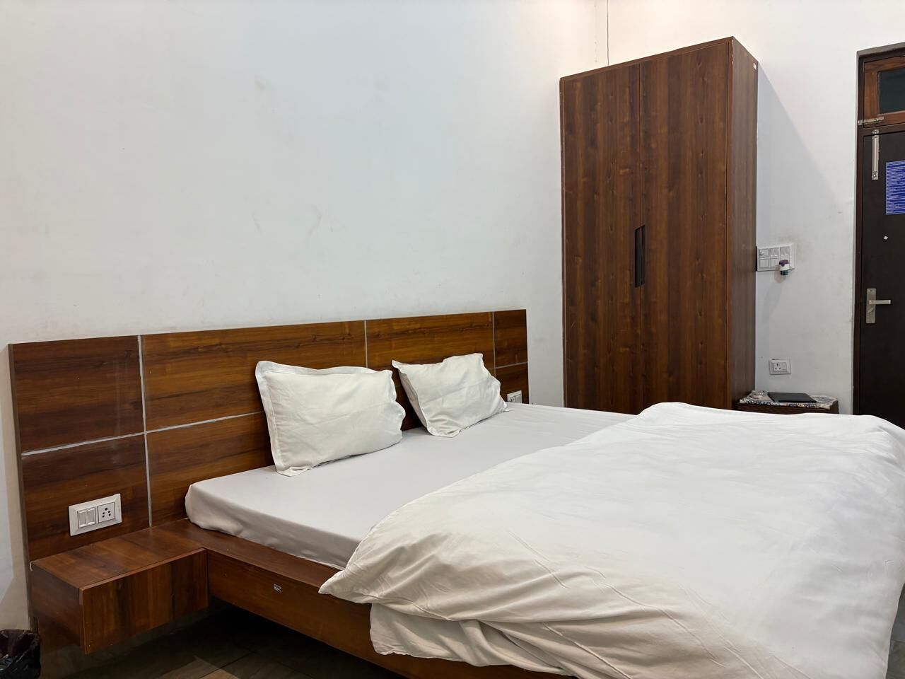 A single bed with a wooden headboard and two pillows, a wooden wardrobe, and a nightstand with a lamp.