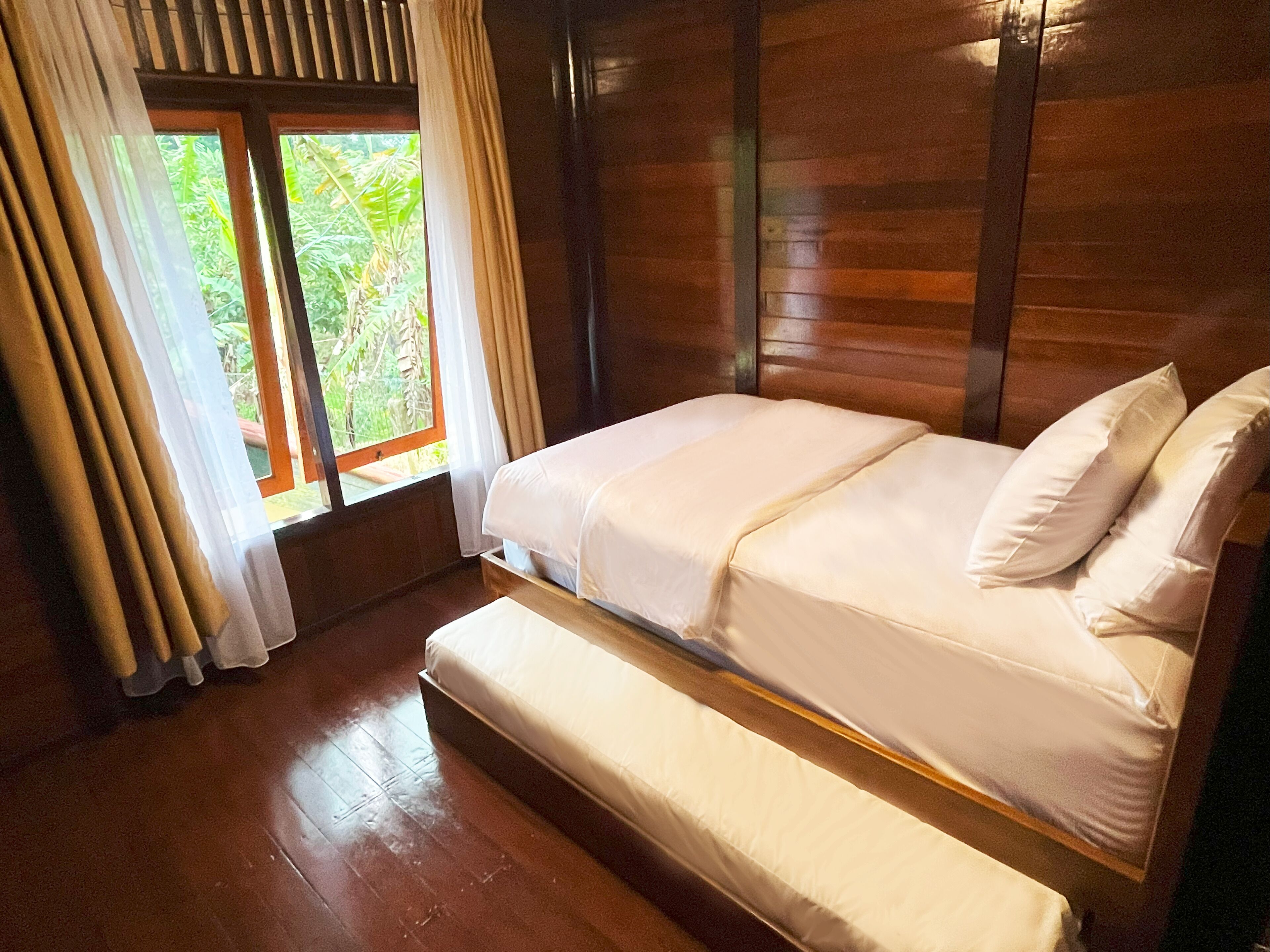 Una cama con estructura de madera y ropa de cama blanca en una habitación con un ventanal y cortinas.