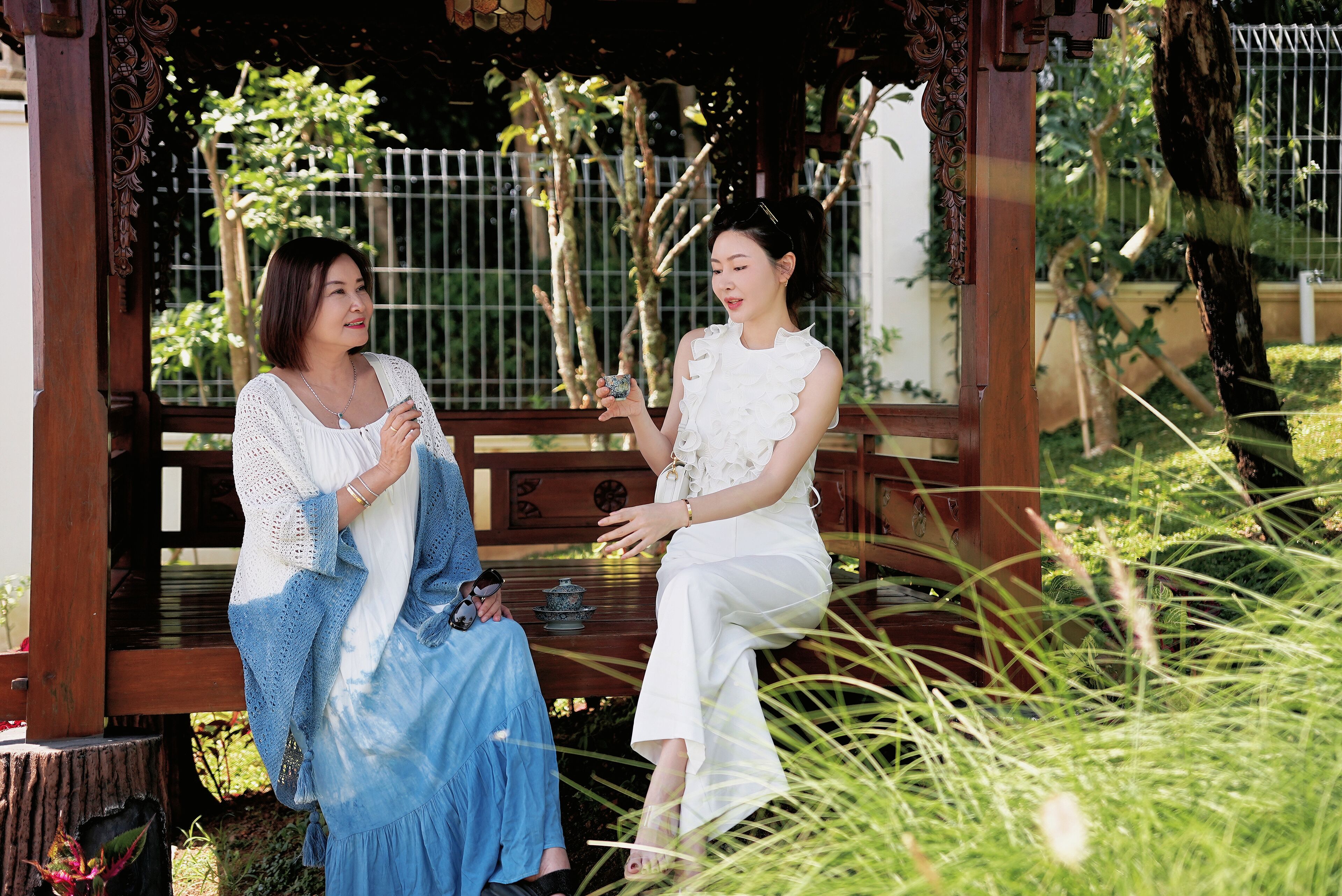 Two individuals seated on a wooden bench in a garden setting with a metal fence and trees in the background.