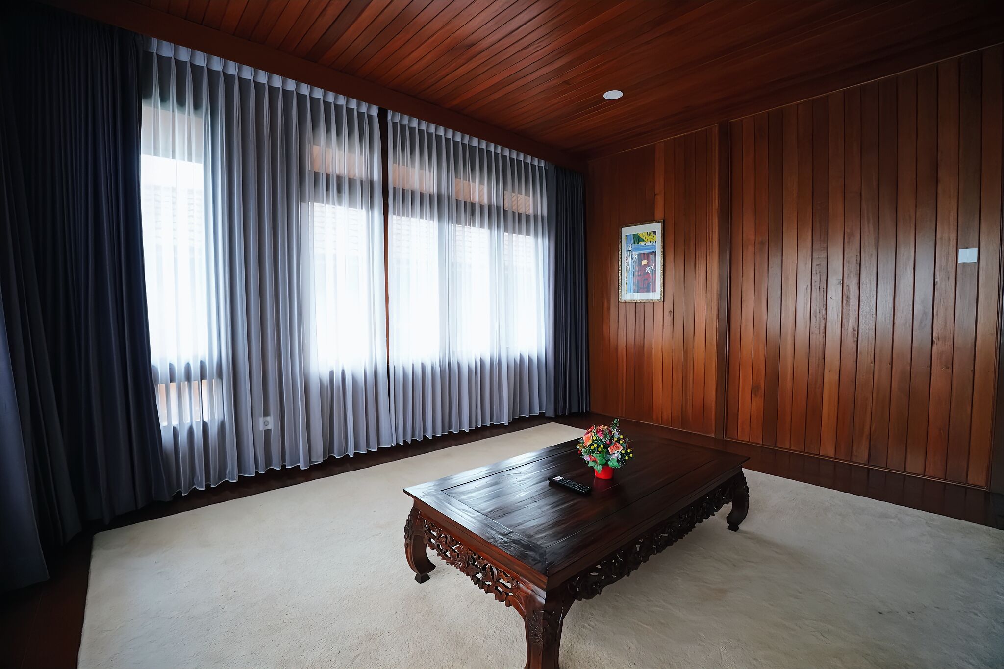 A room with a wooden ceiling, a large window with sheer curtains, a wooden table with a vase of flowers, and a framed picture on the wall.
