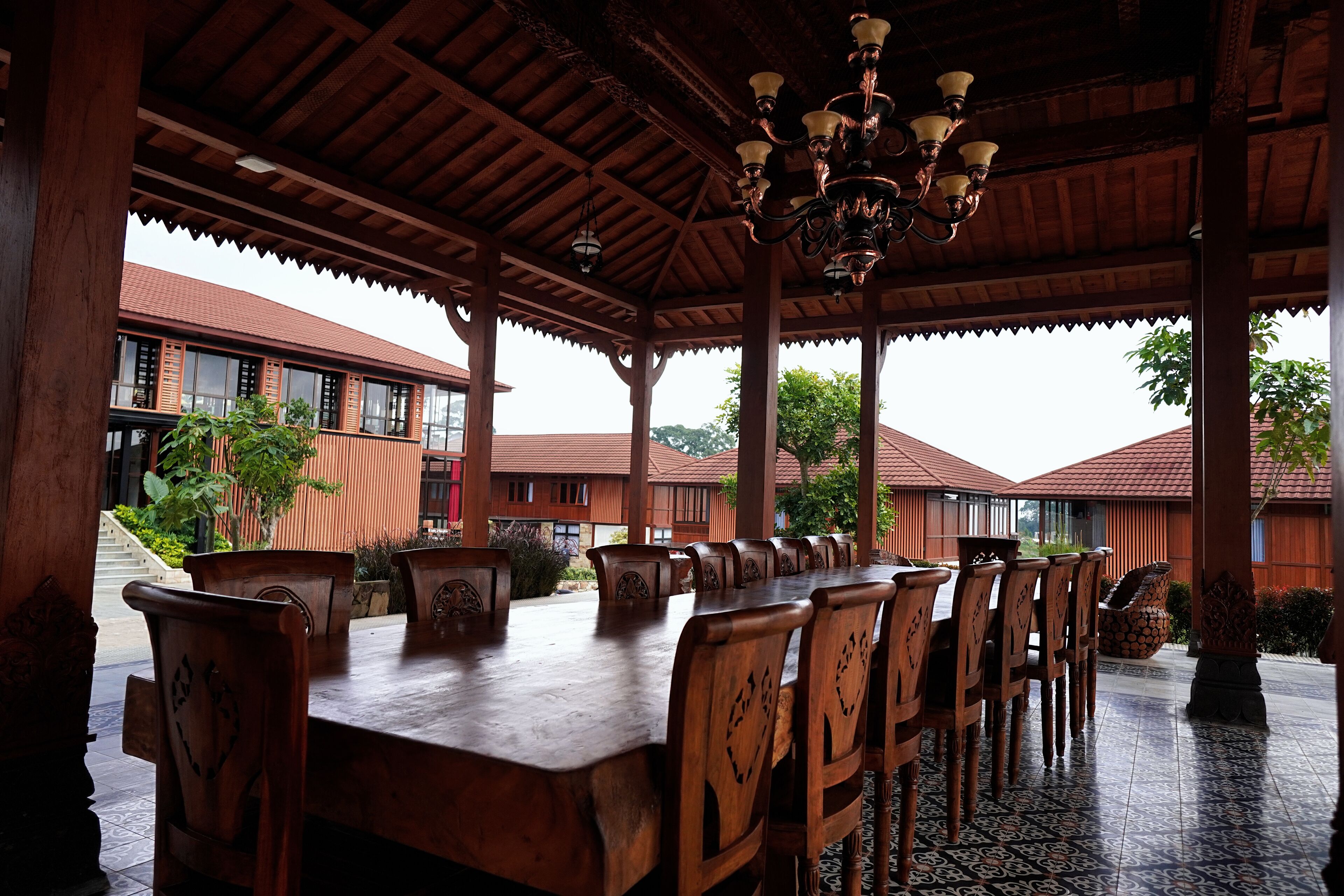 A covered dining area with a long wooden table and chairs, a chandelier, and a view of a building with a red roof.