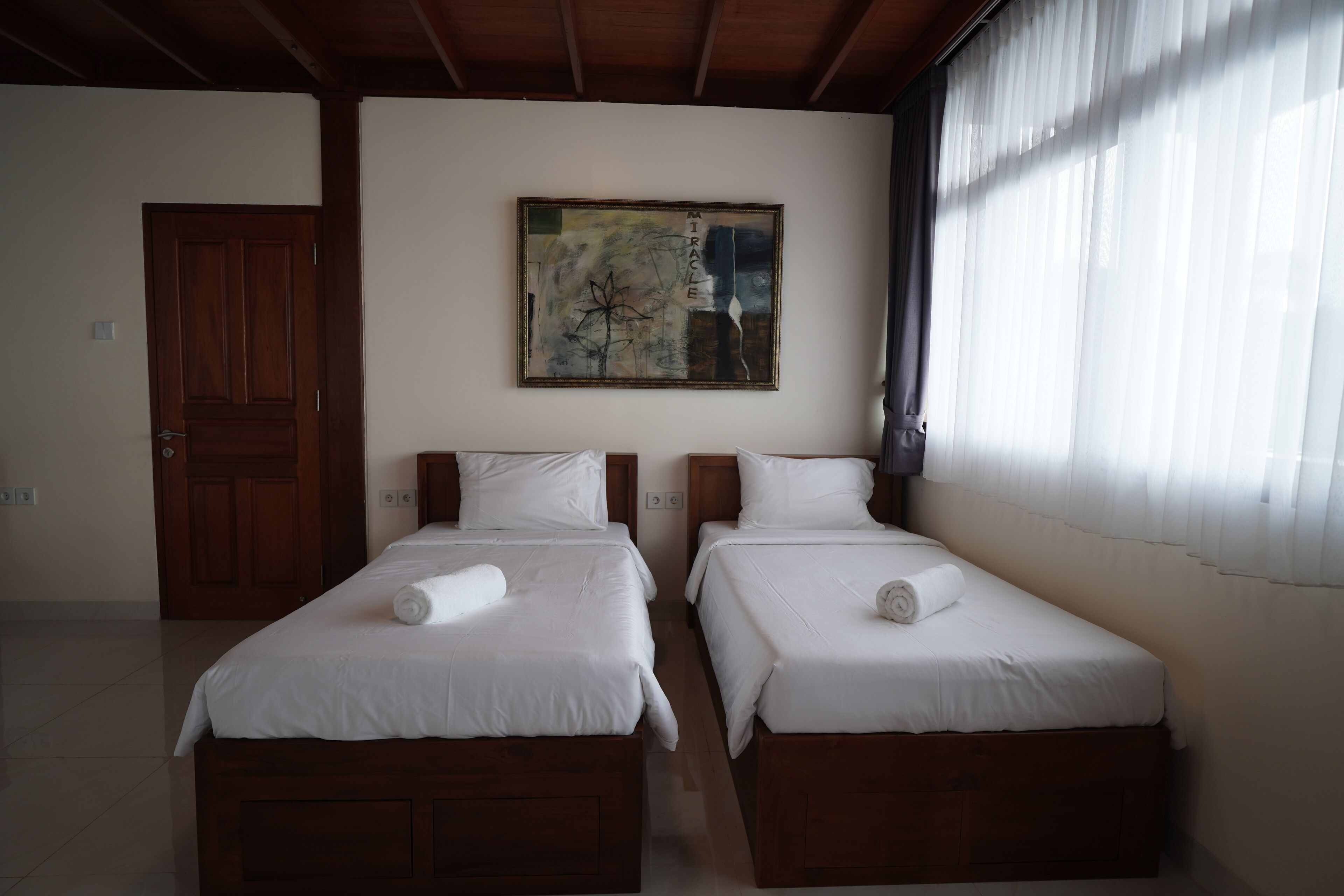 A hotel room with two single beds, a wooden door, a painting on the wall, and a window with curtains.