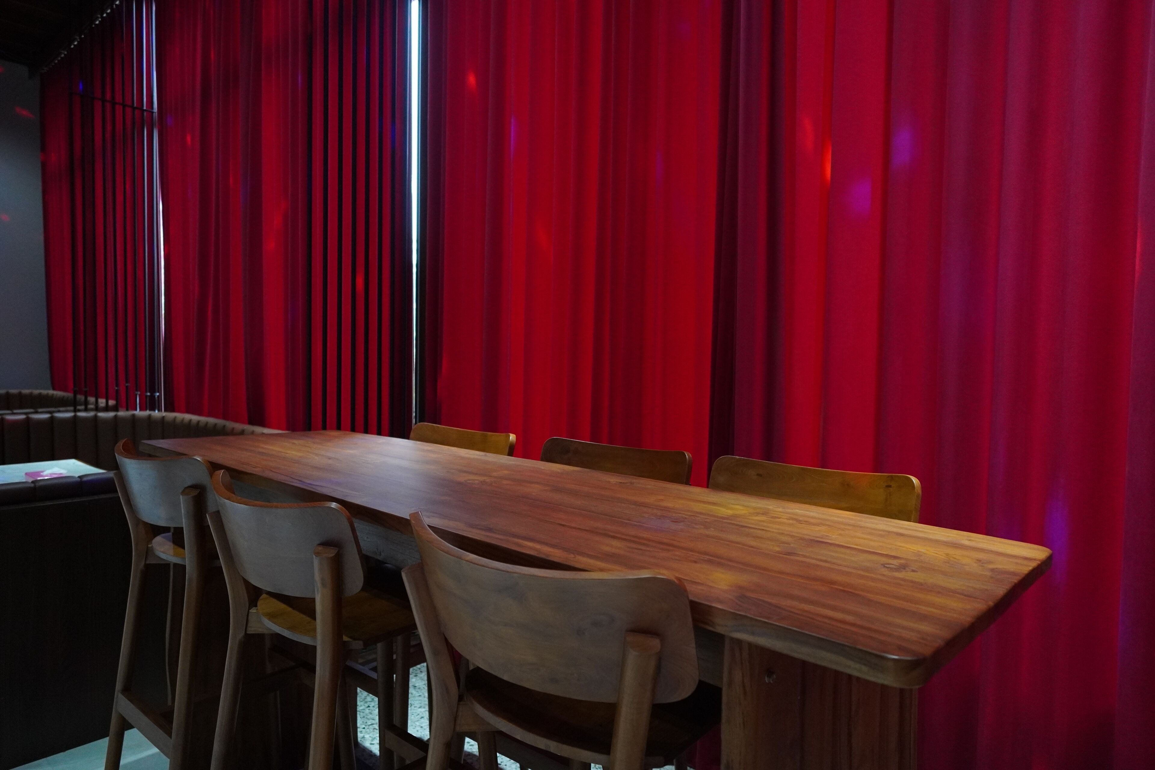 A dining area with a long wooden table and four chairs, set against red curtains.