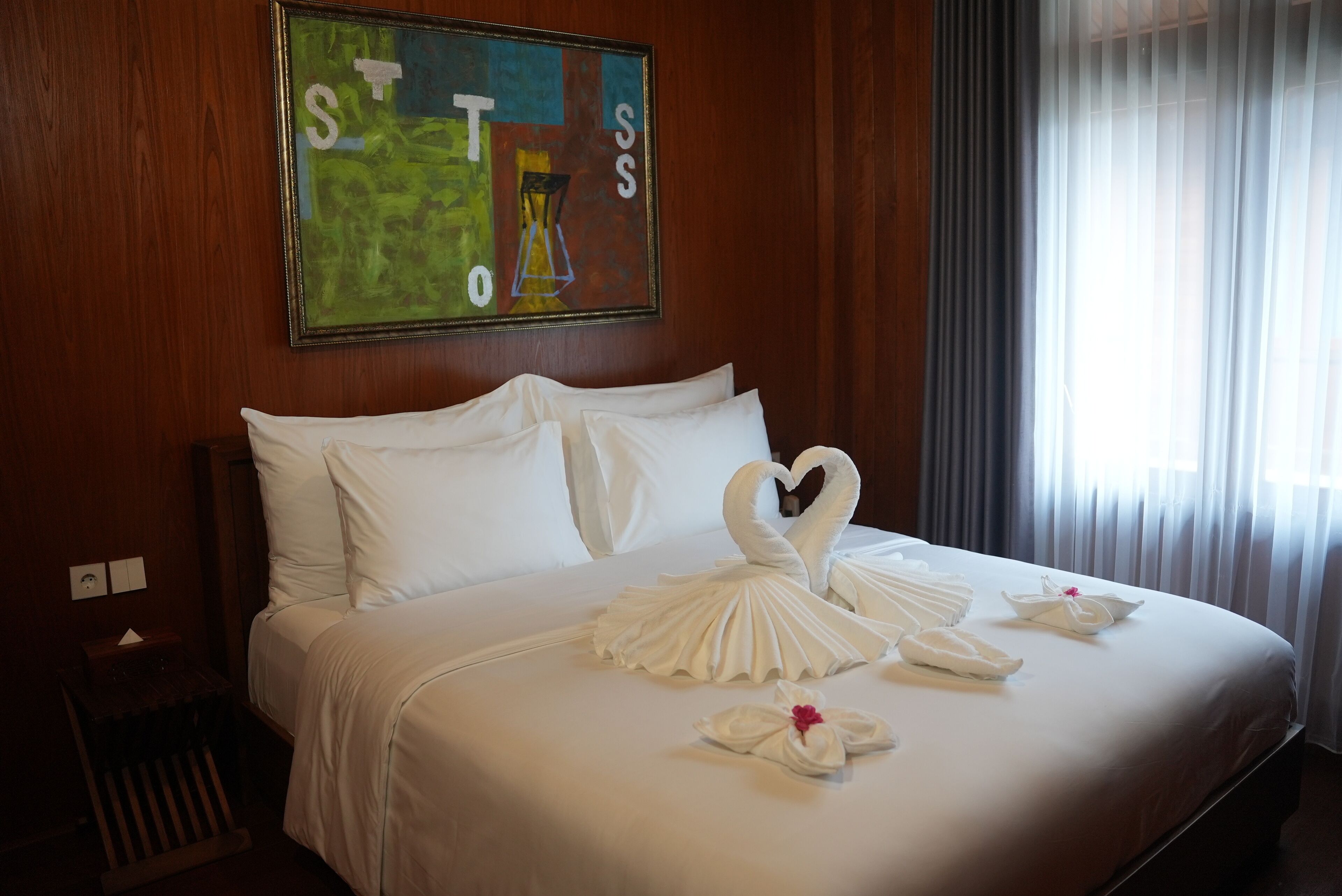 A hotel room with a neatly made bed, a painting on the wall, and a window with curtains.