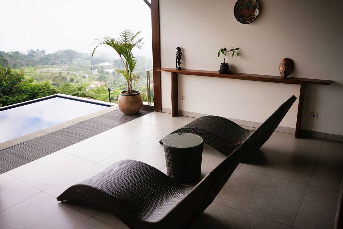 Un espace de détente moderne offrant une vue sur un paysage luxuriant, une étagère en bois agrémentée d’objets décoratifs et une petite table ronde.