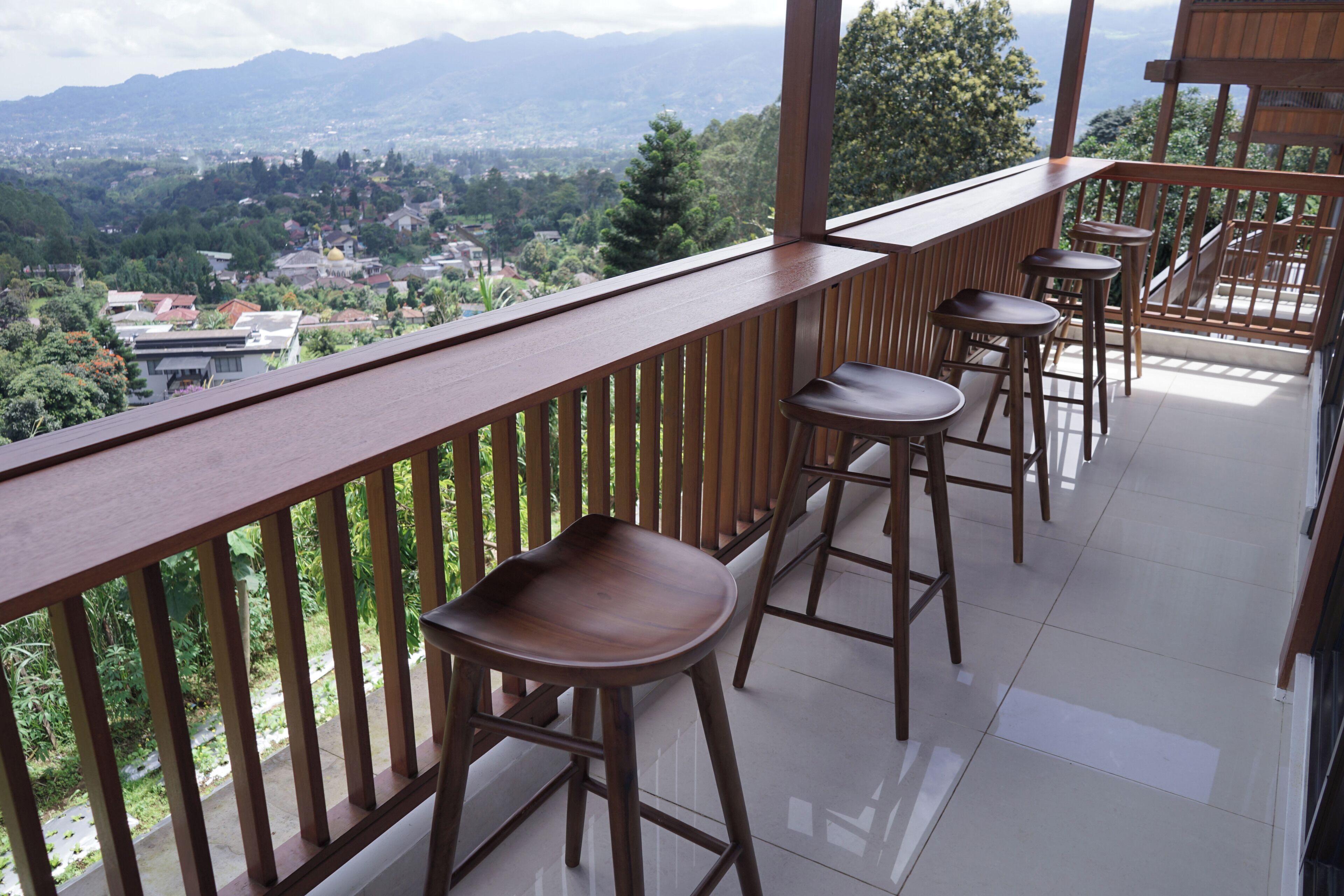 A balcony with a wooden railing, four wooden stools, and a view of a town and mountains in the background.