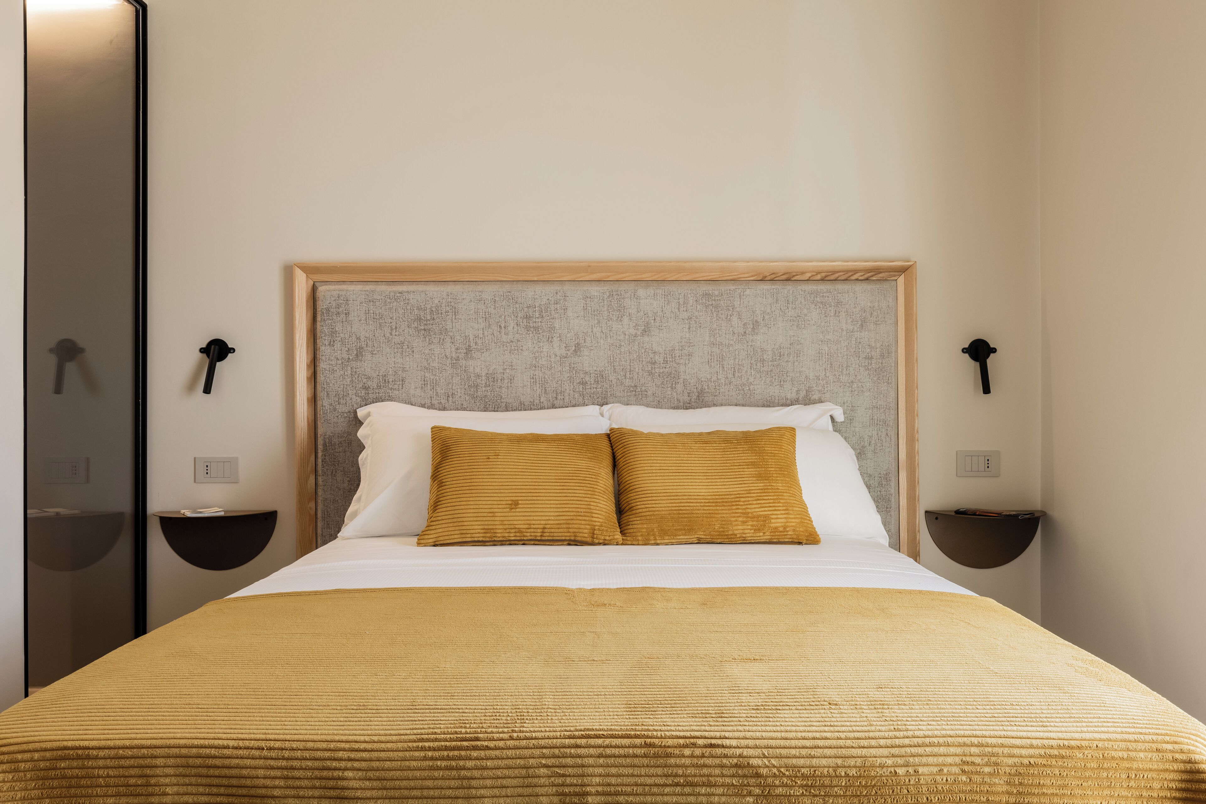 Een netjes opgemaakt bed met een houten hoofdbord, twee nachtkastjes met lampen en een spiegel aan de linker muur.
