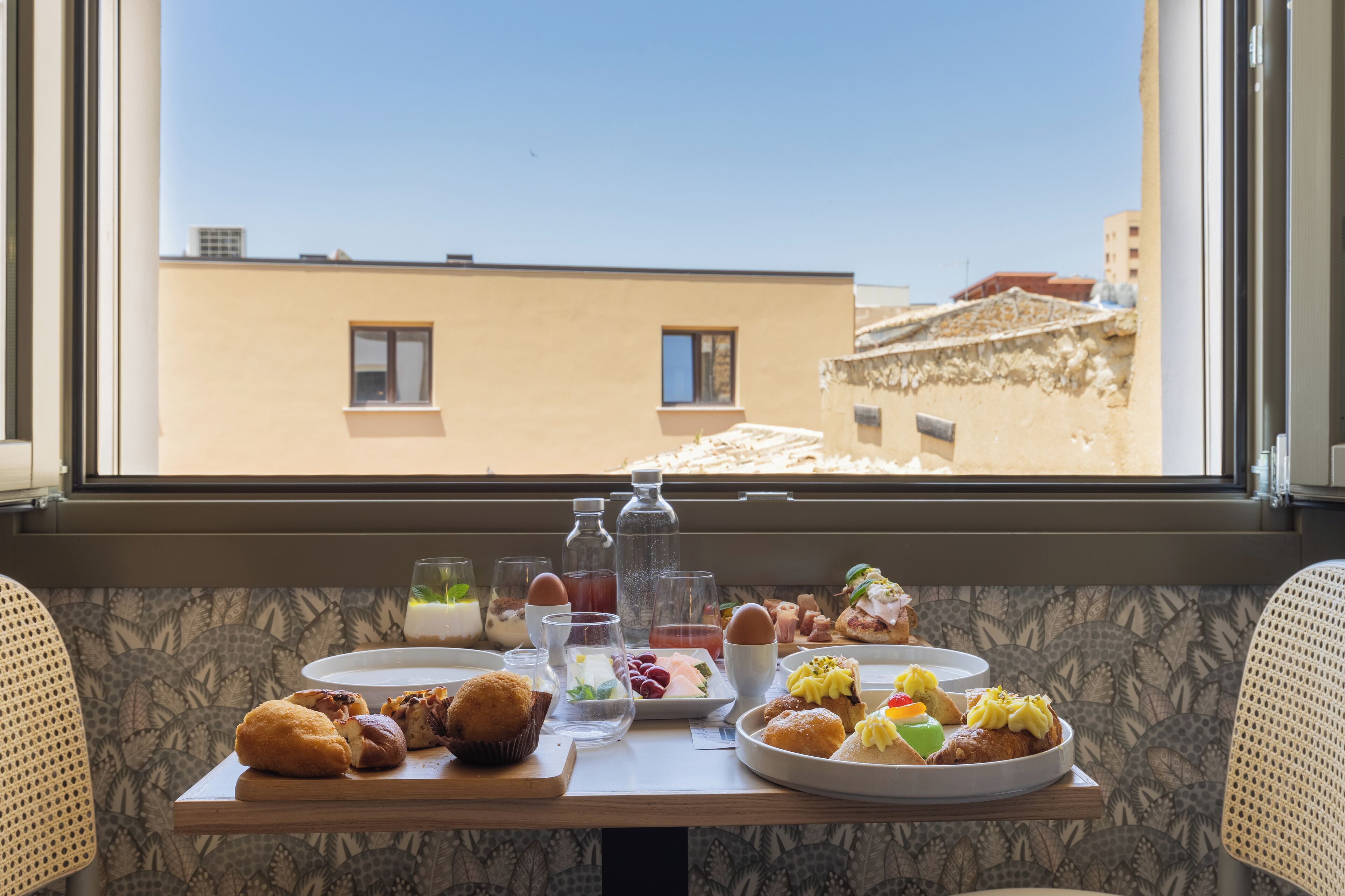A breakfast table with various pastries and drinks, set by a window overlooking a building.