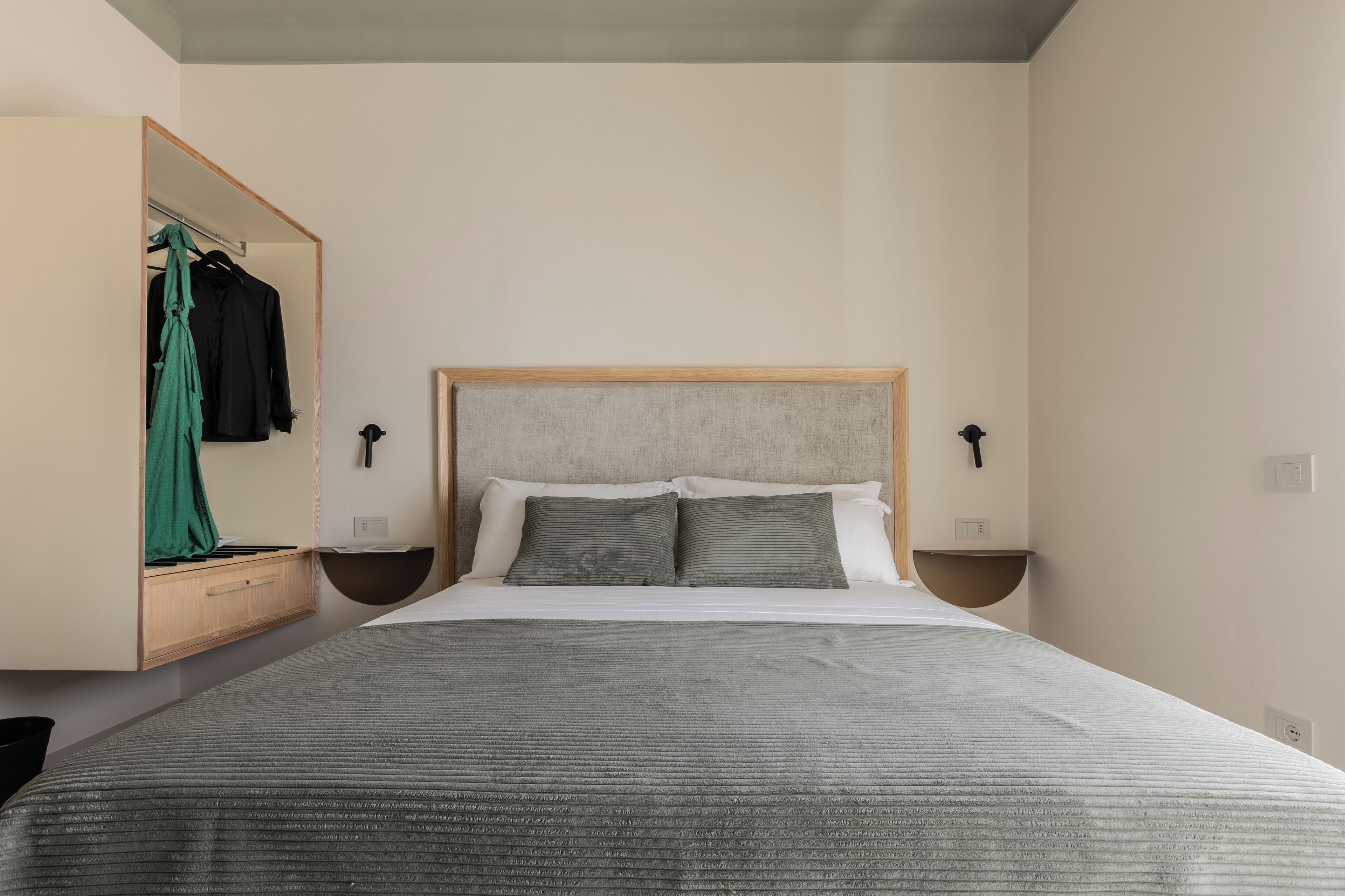 A bedroom with a bed, a wooden headboard, two bedside tables, a mirror, and a hanging green garment.