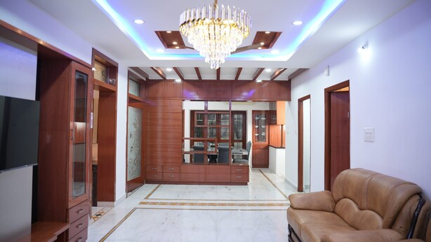 A spacious living room with a chandelier, wooden cabinets, a leather sofa, and a dining area with a glass partition. - Stylish 4-bedroom Family Getaway Luxury Living (Hyderabad)
