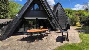 A modern, triangular-roofed house with a patio area featuring a round table and chairs. - Waldnest Eifel - Sauna - Solarium- Pet friendly (Kerschenbach)