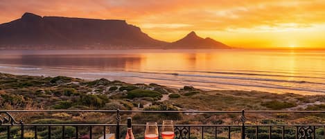 Una mesa con una botella de vino, copas y una tabla de quesos en un balcón con vistas a un paisaje costero con montañas al atardecer.