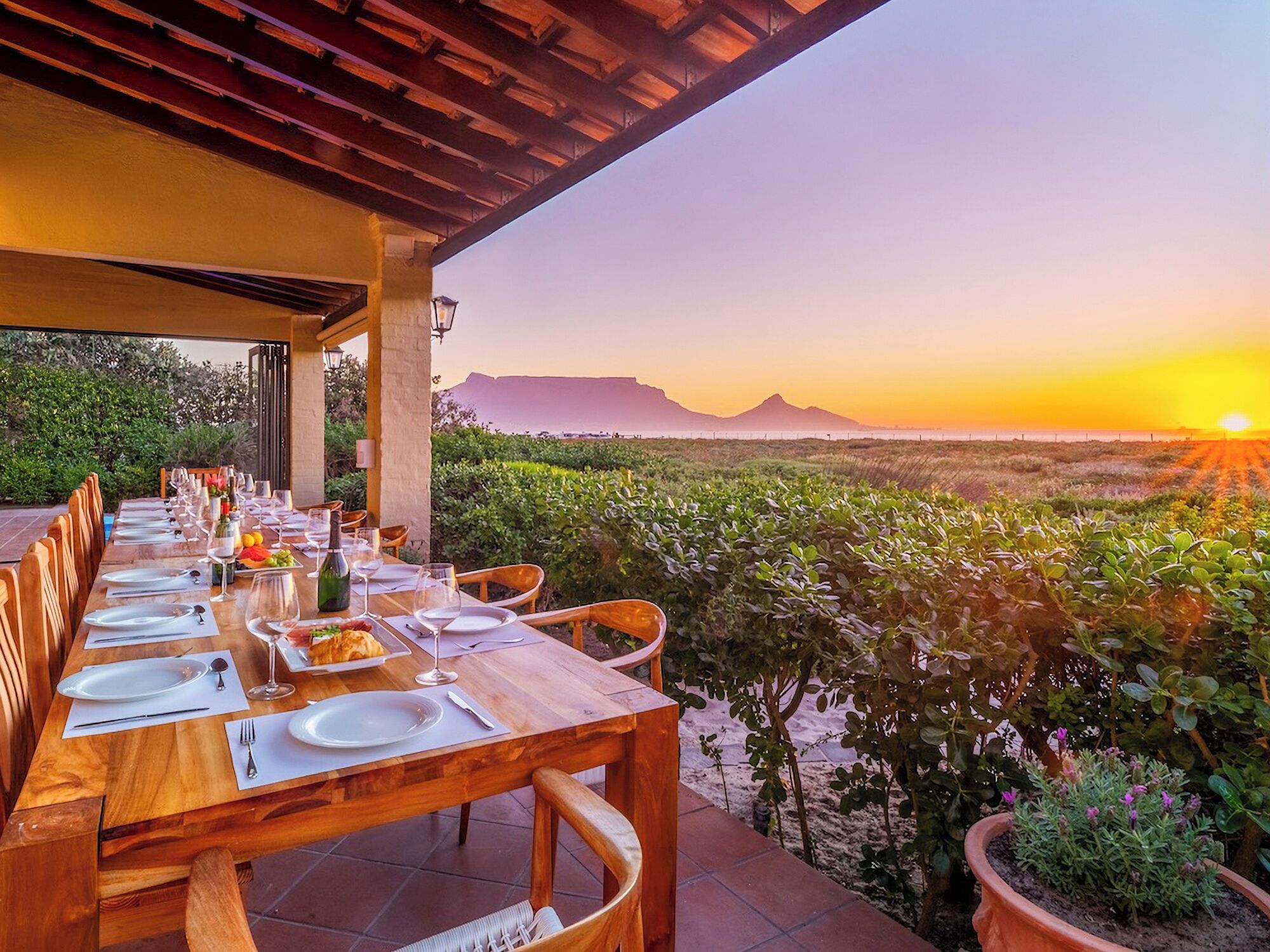 A long wooden dining table set for a meal with a view of a sunset and mountains in the background.