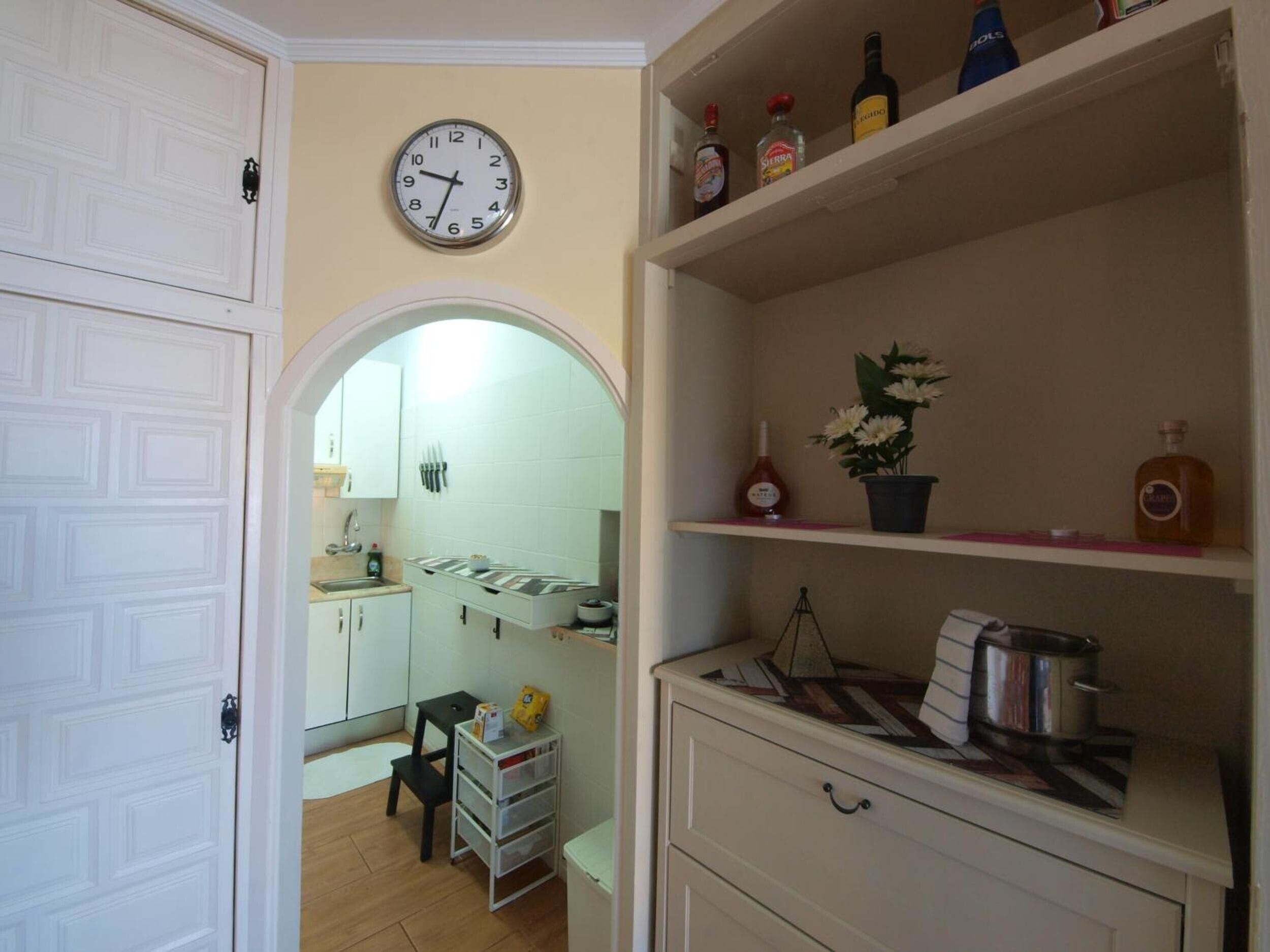 Una cucina con un orologio, scaffali con bottiglie, un lavello e un piccolo tavolo.