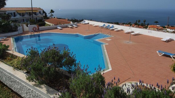 Piscina com vista clara para o mar, rodeada por espreguiçadeiras e um muro branco.