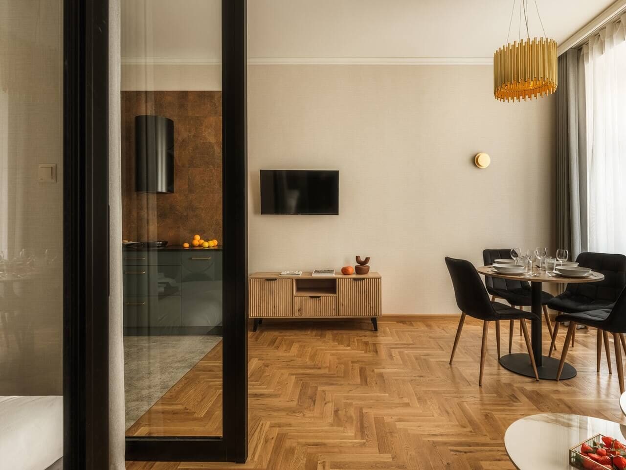 A modern living room with a dining area, a TV mounted on the wall, and a wooden herringbone floor.