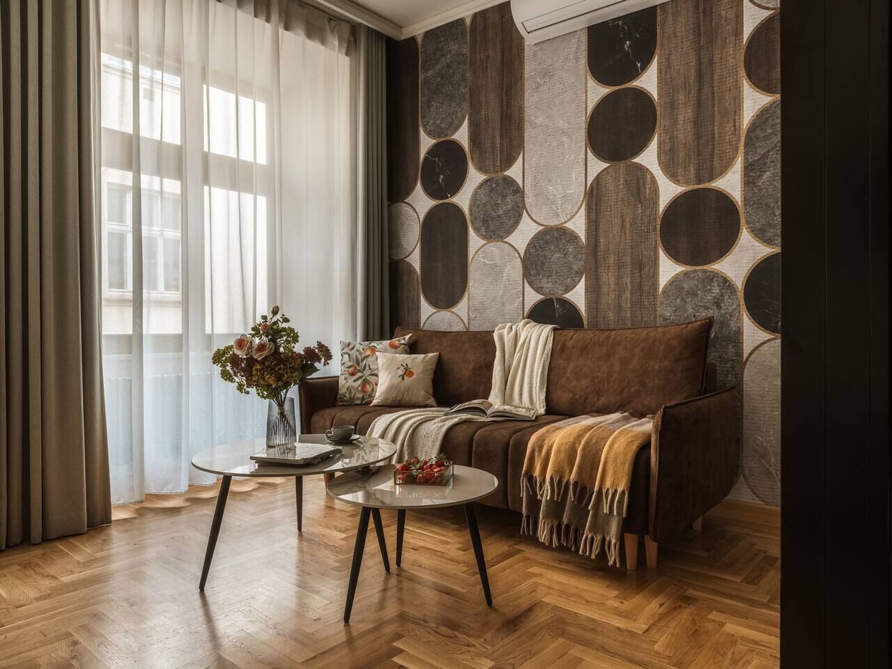 A living room with a brown sofa, two round coffee tables, a vase with flowers, and a patterned wall.