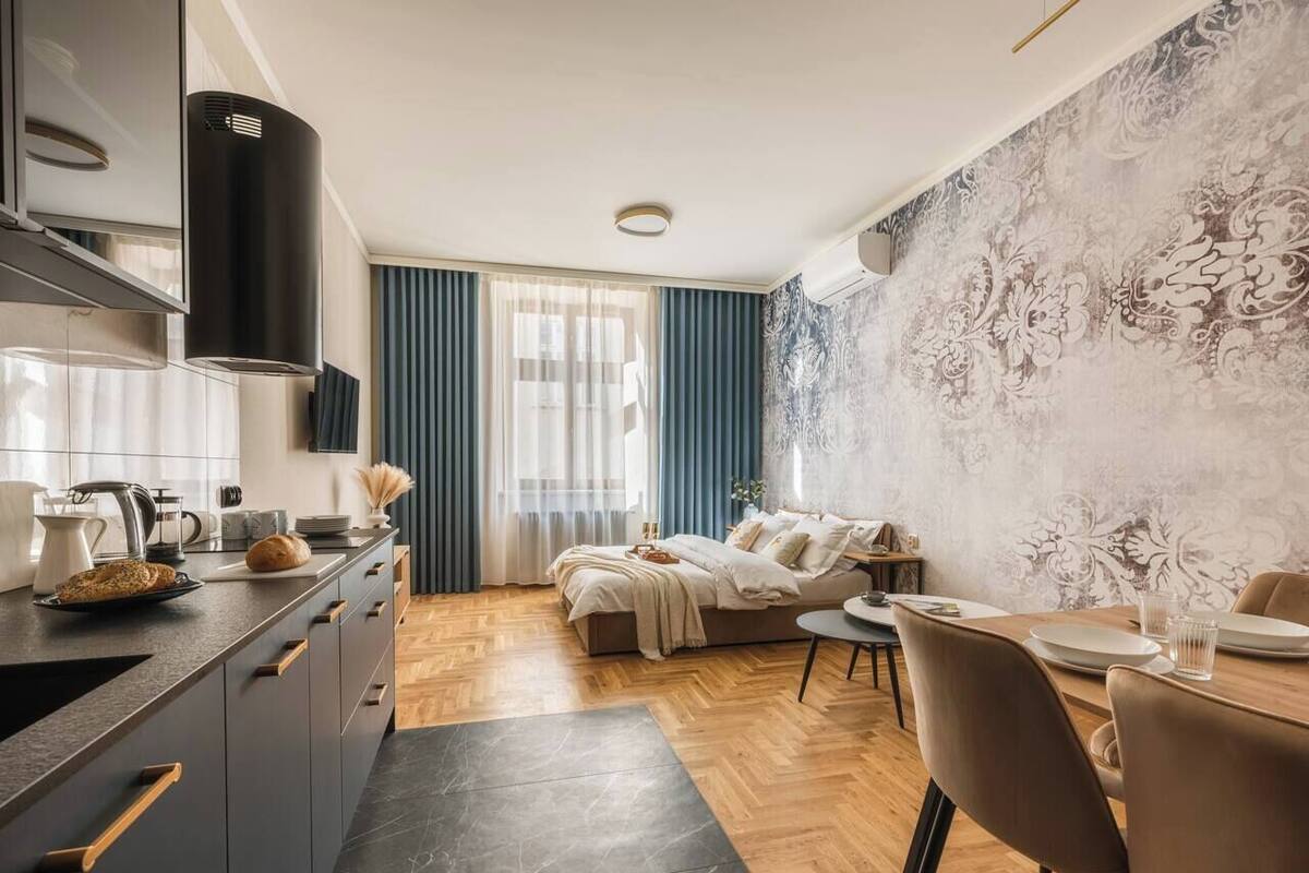 A modern kitchen with a dark countertop, a dining area with a table and chairs, and a bedroom with a bed and curtains.