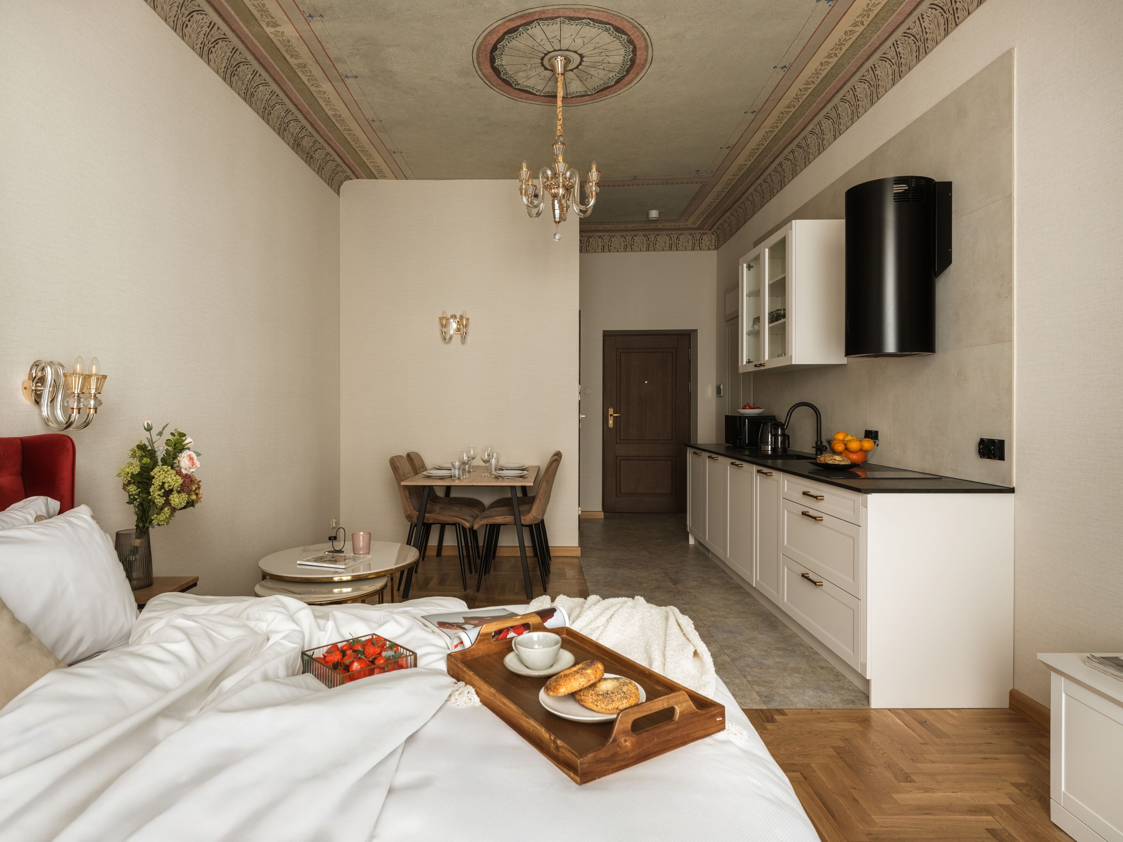 Une chambre avec un lit, une table à manger avec des chaises, une cuisine avec des placards et une cuisinière, et un lustre.
