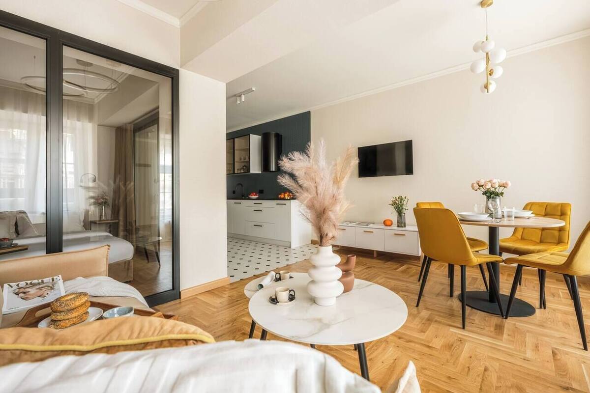 A modern hotel room with a bed, a dining area with yellow chairs, a white table, and a kitchen in the background.