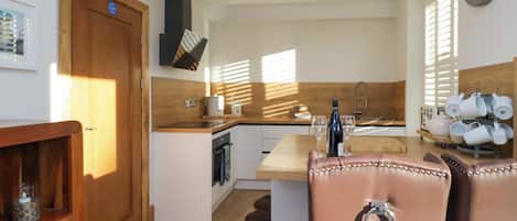 A modern kitchen with wooden cabinetry, a built-in oven, and a large window providing natural light. There is a dining area with leather chairs and a small island with a sink.