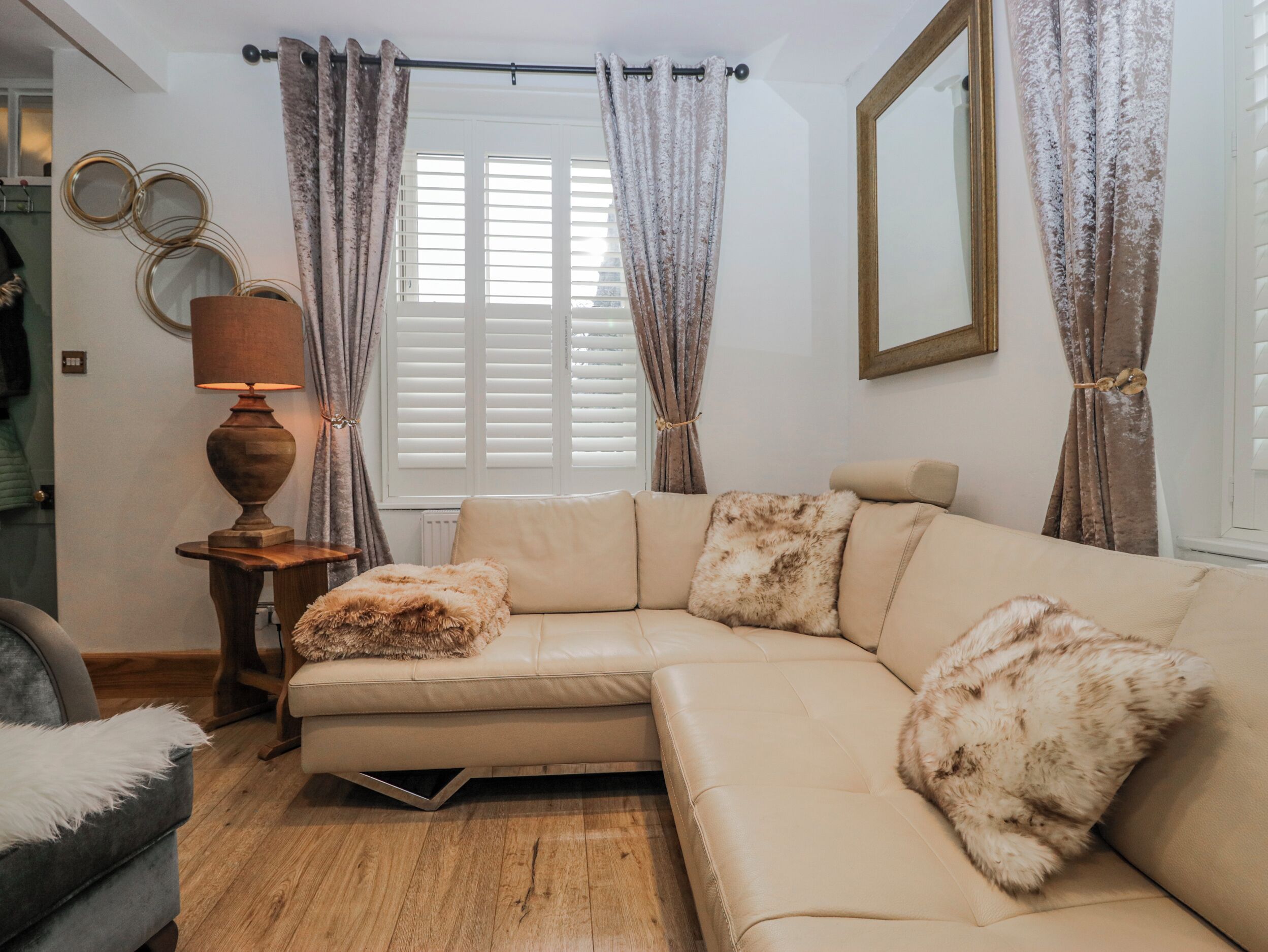Una sala de estar con un sofá seccional color beige, un cojín mullido, una mesita de madera con una lámpara y una ventana con contraventanas blancas.