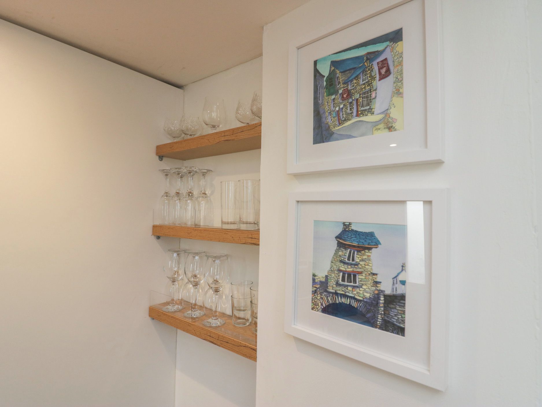 A wall with two framed artworks depicting buildings and a wooden shelf with wine glasses.