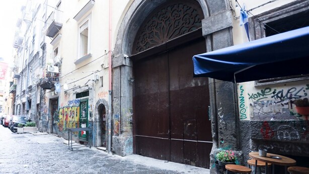 A narrow street with a closed shop with a large wooden door and a blue awning. - GuestHost - Cozy 45 sqm apartment, perfect for 2 people, located on the ground floor of a historic building (1 step to access). A stone's throw from Spaccanapoli (in the restricted traffic zone), the property is ideal for those who want to fully experien (Naples)